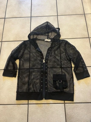 Svart meshjacka med huva H2 Collection storlek S/M - Cool svart meshjacka från H2 Collection med huva, dragkedja och dekorativa öljetter längs öppningen. Jackan har en unik ficka framtill med spänne och ringdetalj. Perfekt för dig som vill sticka ut med en edgy streetstyle-look.