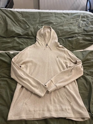 Beige hoodie från Polo Ralph Lauren - Snygg beige hoodie från Polo Ralph Lauren med broderad logga på bröstet. Tillverkad i ett ribbat material med huva och dragsko samt känguruficka framtill. Perfekt för en avslappnad och stilren look.