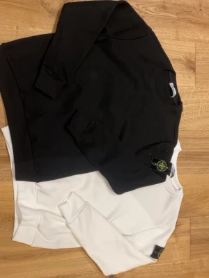 Stone Island svart och vit sweatshirt - Två stilrena sweatshirts från Stone Island, en i svart och en i vitt. Båda har rund hals, långa ärmar och den klassiska Stone Island-loggan på vänster ärm. Materialet är mjukt och bekvämt, perfekt för en avslappnad look. Passar dig som gillar streetwear och enkel design.