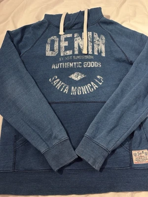 Blå vintage hoodie från Nils Sundström - Snygg blå hoodie från Nils Sundström med tryck på bröstet där det står 'DENIM Santa Monica'. Hoodien har vita dragsnören, känguruficka och ribbade muddar. Perfekt för en avslappnad streetwear-look.storlek M , skit snygg vintage hoddie