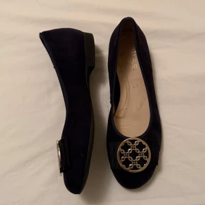 Mörkblå ballerinaskor - Snygga mörkblå ballerinaskor i mocka med dekorativt metallspänne framtill. Skorna har en liten klack men är jättesköna att gå i. Säljer dem då dem inte har kommit till användning och är för små.  Storleken är lite oklar men passar 36-37.