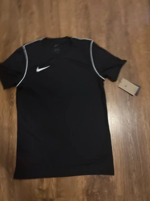 Svart Nike Dri-FIT t-shirt - Snygg svart sport-t-shirt från Nike med vit logga på bröstet och vita detaljer längs axlarna. Tillverkad i Dri-FIT-material som andas och håller dig torr under träningen. Perfekt för gymmet eller löprundan.