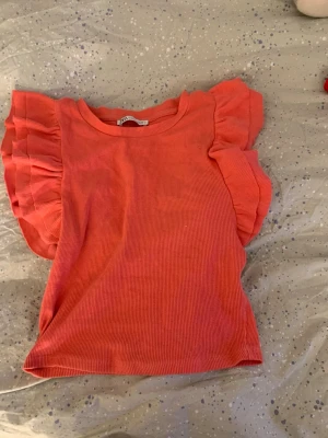 Korallfärgad croppad topp med volangärm - Trendig croppad topp i korall med ribbad struktur och volangdetaljer på ärmarna. Rund halsringning och figurnära passform. Perfekt för dig som vill ha en färgklick i garderoben.