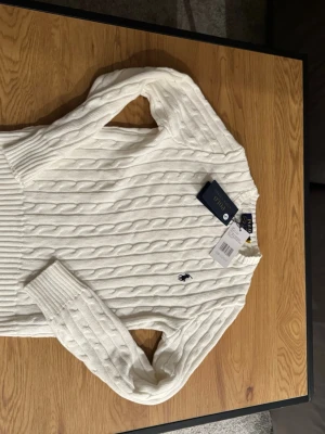 Vit kabelstickad tröja Polo Ralph Lauren - Vit kabelstickad tröja från Polo Ralph Lauren med klassisk broderad logga på bröstet. Tröjan har rund halsringning, långa ärmar och ribbade muddar. Perfekt för en stilren och tidlös look.