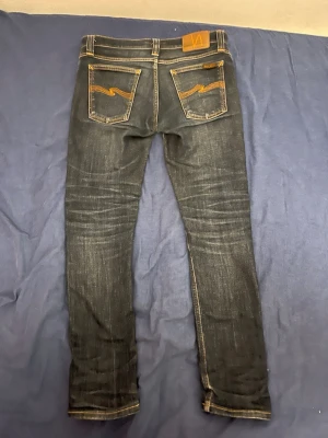 Mörkblåa jeans från Nudie Jeans - Säljer ett par mörkblåa jeans från Nudie Jeans med klassisk femficksdesign och orangea sömmar på bakfickorna. Jeansen har avslappnad passform och är tillverkade i denim med snygga slitningar. Perfekta för en cool och avslappnad stil.