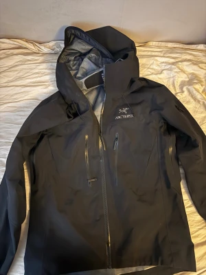 Svart regnjacka från Arc'teryx - Snygg svart regnjacka från Arc'teryx med justerbar huva och vattentäta dragkedjor. Jackan har två bröstfickor med dragkedja och är tillverkad i ett slitstarkt, vattenavvisande material. Perfekt för regniga dagar och outdoor-äventyr.