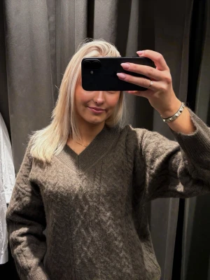 Grön stickad tröja med v-ringning - Mysig grön stickad tröja med v-ringning och diskret kabelstickat mönster. Tröjan har långa ärmar och en avslappnad passform, perfekt för att hålla sig varm under kyliga dagar.