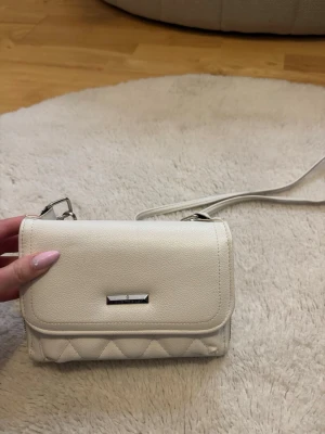 Vit quiltad handväska  - Snygg vit handväska med silverfärgade metalldetaljer. Väskan har en lång axelrem och praktiskt magnetlås. Tillverkad i ett tåligt konstläder med stilren design och diskret logga på framsidan.