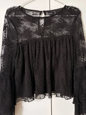 Svart spetsblus  - Aldrig använd! Trendig svart blus i transparent spets från Gina Tricot. Blusen har långa ärmar med volangdetaljer, rund halsringning och knäppning bak i nacken. 
