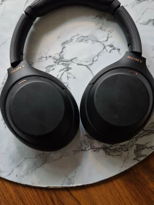 Sony svarta over-ear hörlurar - Stilrena svarta over-ear hörlurar från Sony med mjuka kåpor och diskret logga i guld på båda sidor. Hörlurarna har en modern, rundad design och är tillverkade i plast och konstläder. Perfekta för musik, gaming eller poddar.