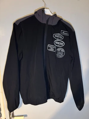 Mercier Vindjacka  - Snygg svart vindjacka från Mercier med grå detaljer på axlarna och flera stilrena Mercier-patchar på bröstet. Jackan är lätt, bekväm och perfekt för vår, sommar eller höst. Dragkedja fram och fickor på sidorna.