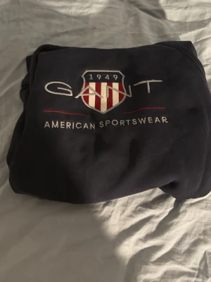 Mörkblå hoodie från GANT barn - Snygg mörkblå hoodie från GANT med broderad logga och texten 'American Sportswear' framtill. Klassisk huva och mjukt material, perfekt för en avslappnad och sportig stil. Sköldmärke med röda och vita ränder ger en retrovibe. Öppen för diskution av pris, barnstorlek