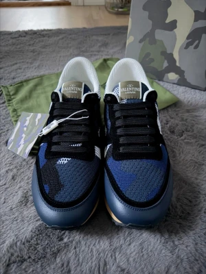 Valentino Garavani blå camo sneakers - Snygga sneakers från Valentino Garavani med blå och svart camo-mönster, svarta skosnören och vit mesh vid hälen. Skorna har en beige sula och detaljer i läder och textil. Perfekta för dig som vill sticka ut med exklusiv streetstyle.