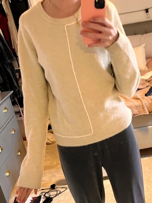 Beige stickad tröja från Zara - Säljer en beige stickad tröja från Zara med rund halsringning och en unik vit söm som går diagonalt framtill. Tröjan har långa ärmar och ribbade muddar vid ärmslut och nederkant. Perfekt för dig som gillar minimalistisk och stilren design.