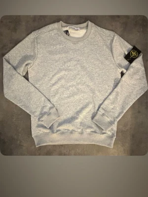 Grå sweatshirt från Stone Island - Snygg grå sweatshirt från Stone Island med klassisk rund hals och ribbade muddar. Tröjan har den ikoniska loggan på vänster ärm och är tillverkad i mjukt bomullsmaterial. Perfekt för en avslappnad och stilren look.