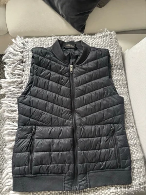 Svart quiltad dunväst med dragkedja - Snygg svart dunväst med quiltat mönster och hel dragkedja framtill. Västen har två fickor med dragkedja och ribbade detaljer vid krage och nederkant. Perfekt för lager på lager och en clean streetstyle-look.