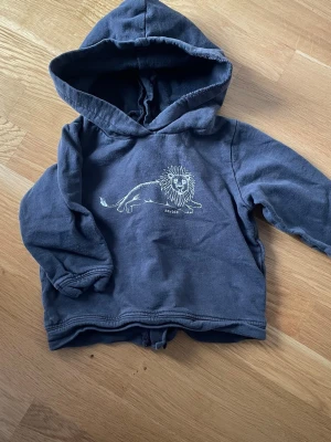 Mörkgrå hoodie med lejon från Newbie 6-9 månader - Mysig mörkgrå hoodie från Newbie med en söt vit lejon-illustration på bröstet. Tröjan har huva och långa ärmar, perfekt för en avslappnad och cool stil. Tillverkad i mjukt bomullsmaterial som känns skönt mot huden. 6-9månadet 