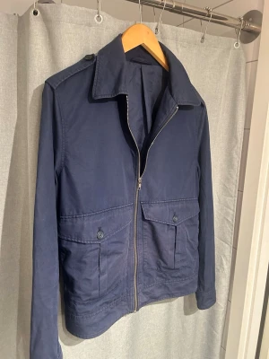 Overshirt blå - Säljer en mörkblå jacka från Filippa K med dragkedja framtill och stora fickor med knappdetaljer. Jackan har en clean och stilren look, perfekt för dig som gillar minimalistisk design. Materialet känns mjukt och är troligtvis bomull. Passformen är normal och ärmarna är långärmade.