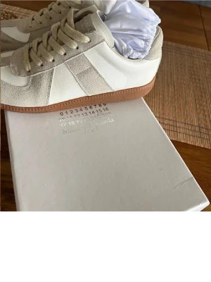 Maison Margiela Replica sneakers beige - Snygga Maison Margiela Replica sneakers i beige och vitt med klassisk gummisula. Ovandelen är i en mix av mocka och läder, med tonade snören och diskreta detaljer. Perfekta för dig som gillar minimalistisk och stilren design.