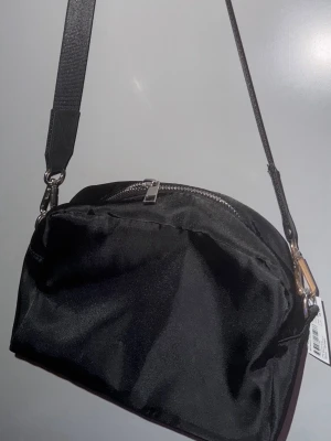 Svart axelväska i nylon - Stilren svart axelväska i glansigt nylonmaterial med justerbar rem och silvrig dragkedja. Väskan har en enkel, rundad form och ett rymligt huvudfack. Perfekt för att bära dina viktigaste grejer när du är på språng.   Från glitter aldrig använd!