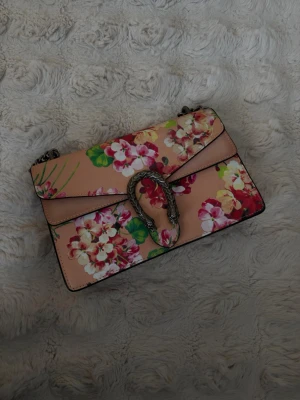 Blommig axelväska med kedja - Snygg axelväska i rosa med färgglatt blommönster och silverfärgad kedjerem. Väskan har ett dekorativt spänne framtill i metall och är tillverkad i ett slätt material. Perfekt för dig som vill sticka ut med en trendig accessoar.