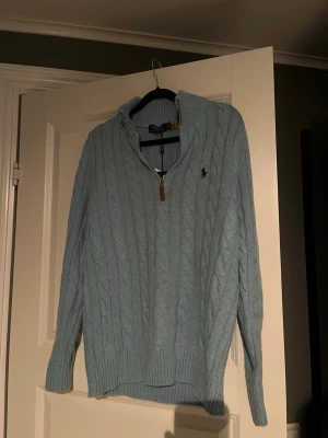 Ljusblå kabelstickad tröja Polo Ralph Lauren - Snygg ljusblå kabelstickad tröja från Polo Ralph Lauren med halv dragkedja och ribbade muddar. Tröjan har en klassisk polokrage och den ikoniska loggan broderad på bröstet. Perfekt för lager på lager och chill stil.