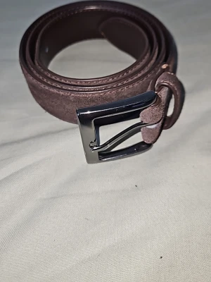 Onewear bälte  - Snyggt brunt bälte i äkta läder från onewear med en robust, fyrkantig metallspänne i silverfärg. Bältet har en klassisk design och passar perfekt till jeans eller chinos. Märket är ingraverat på spännet för en extra cool detalj. 110 cm långt