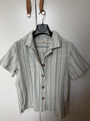Randig kortärmad skjorta  - Kortärmad skjorta från Zara med ljusgrå och vita vertikala ränder. Skjortan har klassisk krage, träknappar framtill och är tillverkad i ett mjukt bomullsliknande material. Perfekt för en avslappnad och stilren look. Köpt i Australien