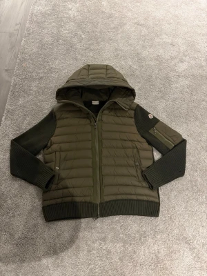 Moncler Cardigan - Snygg olivgrön pufferjacka från Moncler med huva, ribbstickade ärmar och detaljerade fickor. Jackan har dragkedja framtill, två sidofickor och en extra ficka med dragkedja på ärmen. Storlek M skulle nog säga att den kanske också passar nån som har L. 