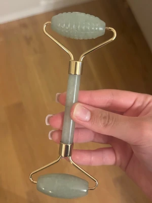 Grön ansiktsroller i jade - Snygg ansiktsroller i grön jade med guldfärgade detaljer. Ena sidan har en slät sten och den andra har en räfflad yta för extra massageeffekt. Perfekt för ansiktsmassage och hudvård. Enkel att använda och ger en lyxig känsla till din rutin.