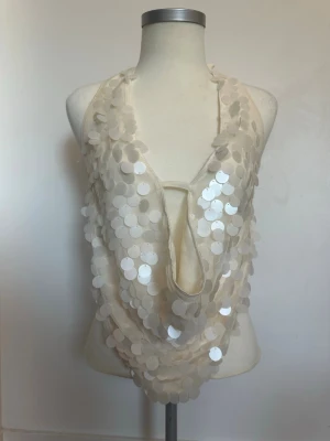Vit paljettprydd halterneck topp - Trendig vit halterneck topp med stora paljetter och djup urringning framtill. Toppen knyts i nacken och har öppen rygg med smal rem. Perfekt för dig som vill sticka ut och glänsa lite extra.