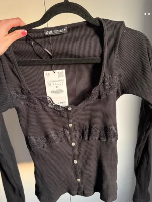 Svart ribbad topp med spets från ZARA - Supersnygg svart ribbad topp från ZARA med spetsdetaljer vid ringningen och tvärs över bröstet. Toppen har v-ringning, knappar framtill och långa ärmar. Perfekt för dig som gillar en feminin och trendig stil. Säljes pågrund av att den tyvärr var lite för liten på mig💗