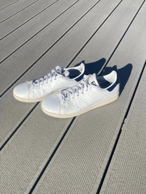 Vita Adidas Stan Smith sneakers - Klassiska vita Adidas Stan Smith sneakers i läder med perforerade ränder på sidorna och mörkblå detalj på hälen. Skorna har vit sula och vita skosnören. Perfekt för dig som gillar en clean och tidlös look.                                      .                                                                                 -Skorna kommer är nytvättade fräscha och har även nya sulor 