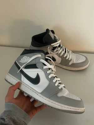 Nike Air Jordan 1 Mid grå/vit - Säljer ett par Nike Air Jordan 1 Mid sneakers i grått och vitt med svarta detaljer. Skorna har klassisk hög siluett, vit tåbox och sula, samt ikoniska Air Jordan-loggan på sidan. Materialet är en mix av läder och syntet. Perfekta för dig som gillar streetwear och snygg stil.