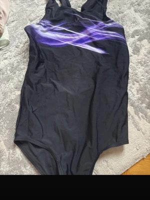 Svart baddräkt från Speedo med lila mönster - Snygg svart baddräkt från Speedo med sportig design och lila/vita grafiska linjer över bröstet. Klassisk modell med breda axelband och rundad halsringning. Tillverkad i stretchigt material som passar perfekt för simning och träning.