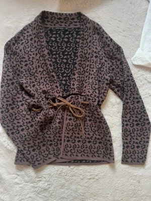 Leopardmönstrad kofta med knytband - Säljer en brun och svart leopardmönstrad kofta från Gina Tricot. Koftan har lång ärm, omlottmodell och ett snyggt knytband i midjan. Perfekt för dig som vill ha en trendig och bekväm look.