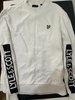 Vit sweatshirt från Lyle & Scott - Vit sweatshirt från Lyle & Scott med relaxed fit. Tröjan har ribbade muddar, rund halsringning och stora svarta logotyper längs båda ärmarna. Klassisk gul logga på bröstet. Perfekt för en avslappnad och sportig stil.