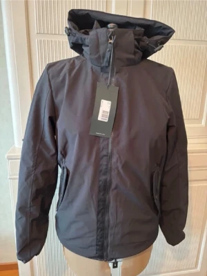 Mörkgrå vindjacka med huva - Snygg mörkgrå vindjacka med justerbar huva och dragkedja framtill. Jackan har två sidofickor med dragkedja och hög krage för extra skydd mot vind. Tillverkad i ett lätt och slitstarkt material som passar perfekt för blåsiga dagar.