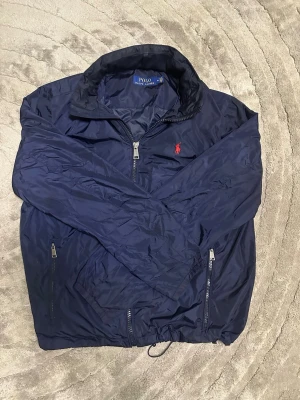 Marinblå vindjacka Polo Ralph Lauren - Snygg marinblå vindjacka från Polo Ralph Lauren med dragkedja, två fickor med dragkedja och justerbar nederkant. Jackan har klassisk krage och röd broderad logga på bröstet. Perfekt för blåsiga dagar och enkel att matcha med olika outfits.