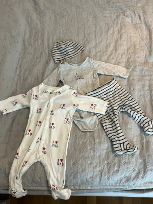 Baby set - Bilden visar ett set med babykläder. Setet inkluderar en body med texten "I love mom", en sparkdräkt med texten "I love dad", ett par randiga byxor och en randig mössa. Kläderna är i ljusa färger och är mysiga.