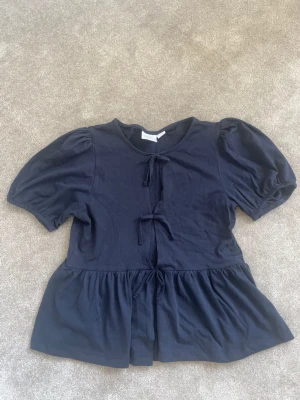 Mörkblå blus med puffärm och rosetter - Supersöt mörkblå blus med puffärmar och volang nedtill. Toppen har två knyt framtill som ger en extra detalj och en lös, flowy passform. Perfekt att styla med jeans eller kjol för en trendig look.