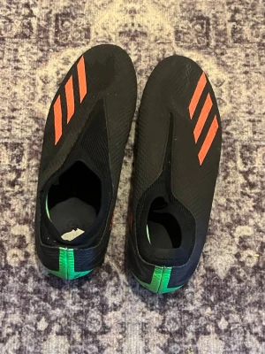 Svarta Adidas fotbollsskor utan snörning - Säljer ett par svarta fotbollsskor från Adidas med klassiska röda ränder på ovansidan och gröna detaljer vid hälen. Skorna har en strömlinjeformad design utan snörning och är tillverkade i syntetmaterial för bästa bollkontroll.