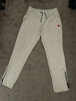 Vita mjukisbyxor från Guess - Säljer ett par vita mjukisbyxor från Guess med diskret logomönster över hela byxan och en röd trekantslogga på låret. Byxorna har svarta dragsnören i midjan och dragkedjor vid bensluten. Perfekta för en chill och stilren look.