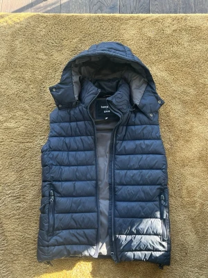 Niche superdry väst - Snygg svart dunväst från Superdry med avtagbar huva och dragkedja framtill. Västen har två fickor med dragkedja och quiltad design som ger en sportig look. Perfekt för lager-på-lager och håller dig varm under kyliga dagar.