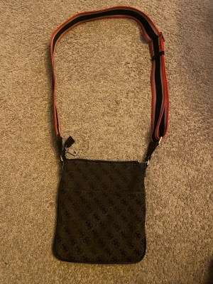 Svart axelväska från Guess - Snygg svart axelväska från Guess med monogrammönster och justerbar axelrem i svart och rött. Väskan har dragkedja upptill och innerfoder med Guess-logga. Perfekt för dig som vill ha en stilren och praktisk väska.