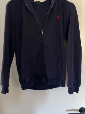 Mörkblå hoodie från Ralph Lauren - Snygg mörkblå hoodie från Ralph Lauren med dragkedja framtill och klassisk röd logga på bröstet. Hoodien har huva, två fickor fram och ribbade muddar vid ärmar och nederkant. Tillverkad i mjukt bomullsmaterial som är skönt att bära. Ny pris 2399kr mitt pris 1750kr. Pris kan diskuteras vid snabb affär.