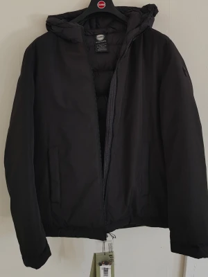 FYND, Colmar svart jacka med luva, storlek L/XL - Svart Colmar dunjacka med huva. Jackan har en dragkedja framtill och två fickor med dragkedja i sidorna. Den är vadderad och har en modern design som passar för olika tillfällen. Jackan är använd en gång och har etiketter kvar.  Storlek 54, motsvarar L/XL med skandinaviska mått  Köpt för 4350:-(ca) på Miinto  Anledning till försäljning är att den är lite kort på mig som är 187 cm. Slutar vid midjan och därför inte använt under hela vintern