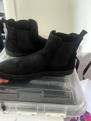 Svarta Chelsea boots i mocka - Säljer ett par helsvarta Chelsea boots i mjuk mocka med elastiska paneler på sidorna och dragflikar bak. Skorna har en chunky sula som ger en modern look och passar perfekt till streetwear eller en clean outfit.