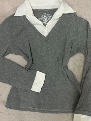 Grå tröja med vit skjortkrage - Snygg grå tröja med fastsydd vit skjortkrage och manschetter för en preppy look. Tröjan har V-ringning och långa ärmar. Perfekt för dig som gillar lager-på-lager-stil utan extra jobb. Materialet känns mjukt och bekvämt. Har ett märkte vid kragen som går bort i tvätten eller med klorin 