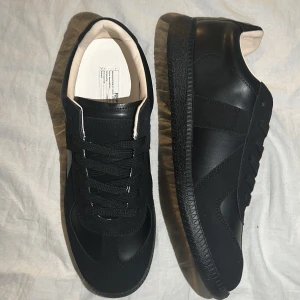 Svarta maison gats - Stilrena svarta sneakers med klassisk låg profil och svart sula. Ovandelen är i en läderliknande finish med matchande svarta skosnören och diskreta sömmar. Insidan är ljus och kontrasterar snyggt mot utsidan. Perfekta för en clean och modern stil.